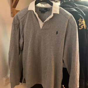 Ralph Lauren Rugby Tröja - Säljer en grå Rugbytröja från RL. Mycket lite använd. Skick 10/10. Vit krage, mörkblå häst. Patches på ärmarna. Storlek M