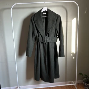 Mörkgrön Kappa  - Fin lång kappa från Gina Tricot premium kollektion. Den är inte så grå som det ser ut som på bilderna utan mörkgrön. Storlek 38 men passar 36-40/42. Går ner till under vaden på mig som är 165. Gott skick, inga defekter.