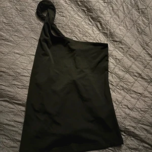 Svart oneshoulder topp från H&M - En elegant svart oneshoulder topp från H&M. Toppen har en asymmetrisk design med en axelbandslös stil på ena sidan och en vriden axelrem på den andra. Perfekt för festliga tillfällen samt vardagligt. använd 1 gång