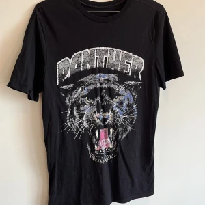 Svart t-shirt med pantertryck - En svart t-shirt med ett stort tryck av en panter och texten 'PANTHER' på framsidan. T-shirten har korta ärmar och är tillverkad av 100% bomull.