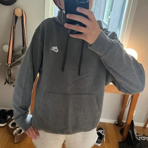 Grå hoodie från Nike - En bekväm grå hoodie från Nike med en vit logotyp på bröstet. Hoodien har en känguruficka och svarta dragsnören vid huvan. Den har långa ärmar och ribbstickade muddar vid ärmslut och nederkant.
