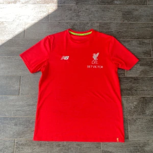 Röd fotbollströja från New Balance - En röd fotbollströja från New Balance med korta ärmar. Tröjan har LFC-logotypen och BetVictor som sponsor på framsidan. Den har även New Balance-logotypen på bröstet och en liten LFC-logotyp på nederkanten.
