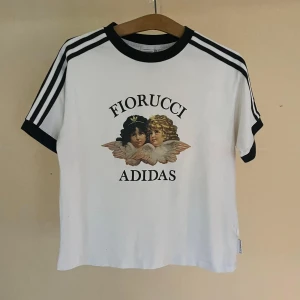 Vit t-shirt från Adidas och Fiorucci - En vit t-shirt från Adidas och Fiorucci med ett tryck av två änglar på framsidan. T-shirten har svarta detaljer vid kragen och ärmsluten samt svarta ränder längs ärmarna.
