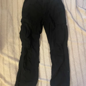 Svarta lowrise lågmidjade y2k cargobyxor - Svarta lågmidjade cargobyxor från Brandit Storlek W30 passar S/36 Finns lite slitage på botten av benen men tycker att det adderar till den tuffa looken