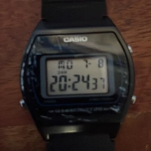 Svart digital klocka från Casio - En svart digital klocka från Casio med modellnummer W-202. Klockan har en rektangulär urtavla med digital display som visar tid, datum och dag. Den har en svart plastrem och en baksida i rostfritt stål. Klockan är vattentålig upp till 50 meter. Lite repig 