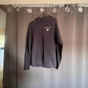 Svart hoodie från GANT - En svart hoodie från GANT med en broderad logotyp och ett emblem på bröstet. Hoodien har en känguruficka framtill. passar storleken S/M