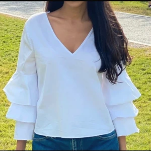 Vit blus från Zara Basic - En elegant vit blus från Zara med v-ringning och långa ärmar. Blusen har volangdetaljer på ärmarna och en öppen rygg med en horisontell rem.