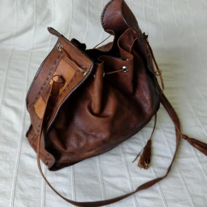 Vintage läderväska - brun rymlig axelremsväska  - Skitfin vintage väska i läder. Den är ca 34x40 cm stor och remmen är ca 105 cm lång. Den är i fint skick (ett par små fläckar i fodertyget och ett litet läderband har lossnat i en detalj i ena sidan). 