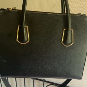 Handväska - Svart handväska med gulddetaljer. Säljer då den inte kommer till användning. Väskan har några synliga skador (se bilder), därav priset. Nypris 400💓