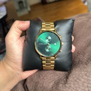 Guldfärgad klocka från Alfred Sung - En helt ny o elegant klocka från Alfred Sung med guldfärgad metallarmband och grön urtavla. Klockan har en datumvisare och två mindre urtavlor för extra funktioner. Den levereras i en svart förpackning med Alfred Sung-logotypen.