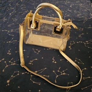 Genomskinlig väska - Genomskinlig väska med guldiga detaljer och en avtagbar rem. Mått är ungefär B=27cm D=13cm H=20cm. Lite ihoptryckt på bilden, men det går att töja ut den med lite saker i.