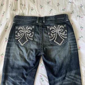 Jeans med detaljerade bakfickor  - Snygga raka jeans med detaljer ifrån Affliction i storlek 36 (ca storlek L). Midjemåttet tvärsöver är 48cm tvärsöver (ca 96 runtom hela) och innerbenslängden är 74cm långt. Nypris: Runt 1500kr