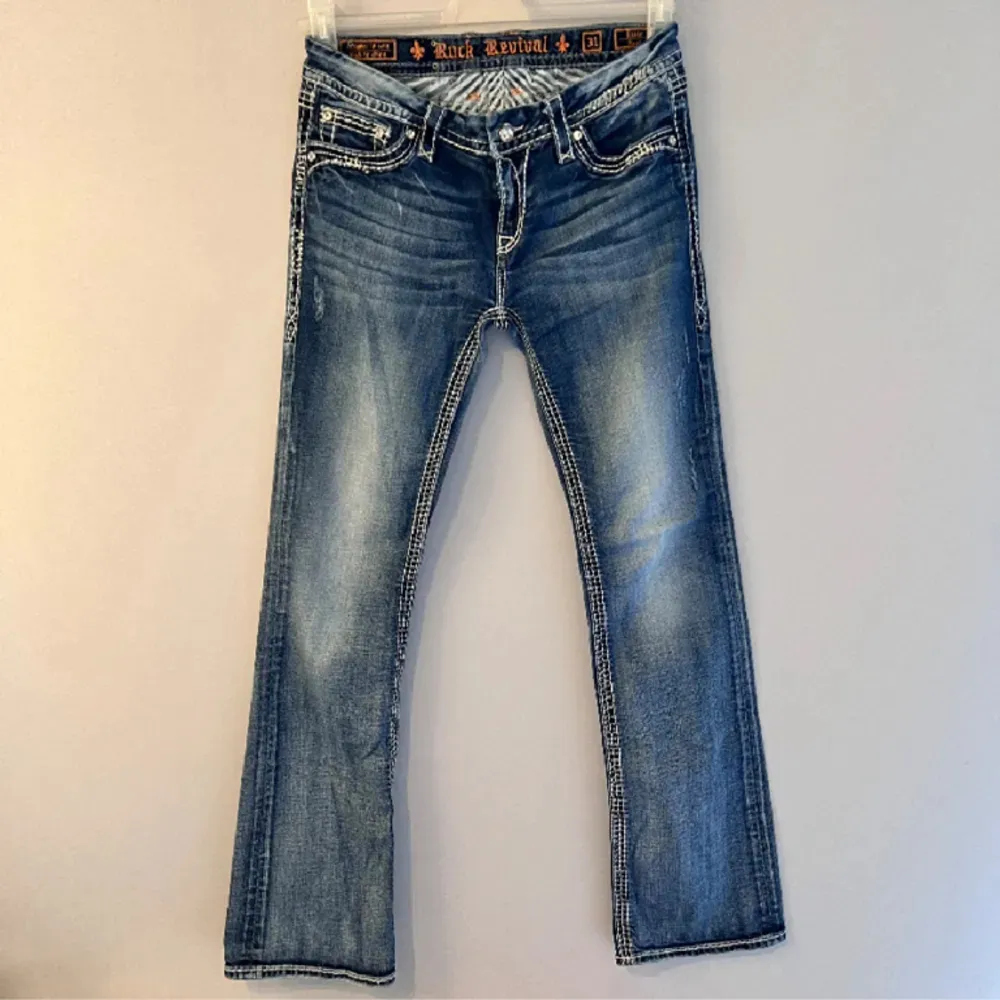 Rock Revival bootcut jeans. Vita designstygn både fram och bak med kristallnitar. Bra skick på tyget men lite nitar som lossnat och framknappen har en spricka (enligt bilderna). Nypris: Rock Revival bootcut ca 1900kr. Hör gärna av dig om du har frågo. Jeans & Byxor.