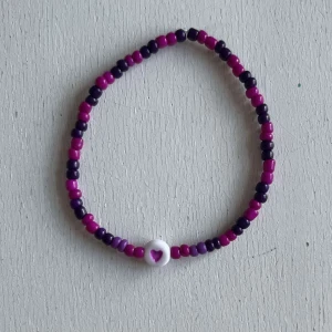 Egengjort armband - Ett egengjort pärlarmband med färgerna magenta, mörklila och en ännu mörkare lila. Armbandet har en annan pärla i vit som har en rosa hjärtsymbol på. 