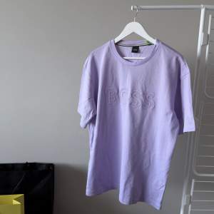 Nyköpt från sommarkollektionen av Hugo Boss en pastell lila Teecomfort T-shirt med BOSS print. Oanvänd, Nypris 1045 kr. Material: 100% Bomull. Storlek: XL