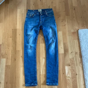 Blå jeans från Morris - Säljer ett par snygga blå jeans från Morris i storlek 31/32. De har en klassisk femficksdesign med en liten sliten detalj vid bältesslingan och en liten reva vid bakfickan. Jeansen är i bra skick men har några slitningar som ger dem en cool look. Perfekta för vardagsbruk!