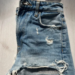 Blå jeansshorts från Zara - Säljer ett par snygga blå jeansshorts från Zara i storlek XS. De har slitningar och fransar. Perfekta för sommaren och ger en avslappnad och trendig stil. Shortsen har en normal passform och är tillverkade i bomull. Använda ett fåtal gånger därför inte så slitna.