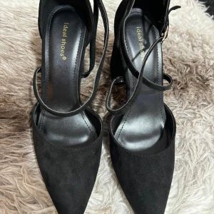 Svarta pumps från Ideal Shoes - Säljer ett par eleganta svarta pumps från Ideal Shoes. De har en spetsig tå och en bekväm klack. Skorna är gjorda av mocka och har en snygg remdesign som ger extra stöd och stil. Perfekta för fest eller andra speciella tillfällen!