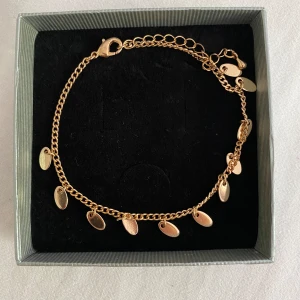 Guldarmband med små hängande berlocker - Säljer en superfin guldfärgad fotlänk med små hängande berlocker. Fotlänken är i kedjestil och har en justerbar längd med en karbinhake som lås. Perfekt för att ge en extra touch till din outfit, oavsett om det är till vardags eller fest! 