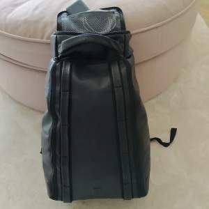 Svart ryggsäck - En svart ryggsäck med flera fack och dragkedjor. Ryggsäcken har vadderade axelremmar och en hård baksida för extra stöd. Inuti finns nätfickor för organisering av småsaker. Märket DB.