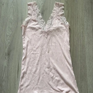 Ljusrosa linne med spetsdetaljer - Ett ljusrosa linne med spetsdetaljer runt halsringningen och axlarna. Linnet är ärmlöst och har en v-ringad design.