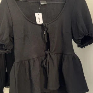 Svart knytblus från Gina Tricot - Säljer en snygg svart blus från Gina Tricot. Blusen har korta ärmar med volangdetaljer och en knytning framtill. Perfekt för både vardag och fest! 🖤 Helt ny, skriv för frågor!