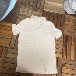 Beige kortärmad t-shirt från Dressmann - Säljer en beige kortärmad t-shirt från Dressmann i storlek S. T-shirten är i bra skick och perfekt för vardagsbruk. Den har en klassisk design med en liten krage och knappar vid halsen. Materialet känns mjukt och bekvämt.