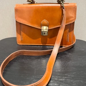 Brun retroväska i äkta skinn, Vera Pelle - Superfin väska i äkta skinn som kan bäras antingen på axeln eller i handen. Axelremmen är avtagbar. Väskan har ett låsspänne som man kan låsa med en nyckel som medföljer. Väskan är i mycket fint skick, 2 små streck som blir naturligt av användning. 