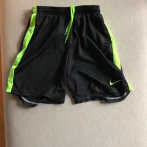 Svarta träningsshorts från Nike - Svarta träningsshorts från Nike med gröna detaljer och dragsko i midjan. Tillverkade i Dri-Fit material för att hålla dig torr och bekväm under träning.