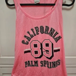 Rosa linne från Gina Tricot - Ett rosa linne från Gina Tricot med texten 'California 89 Palm Springs' tryckt på framsidan. Linnet har en rund halsringning och är ärmlöst.