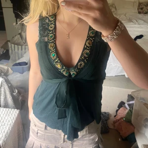 Mörkgrön topp med pärldetaljer - En mörkgrön topp med pärldetaljer runt halsringningen. Toppen har en knytning framtill och är ärmlös.