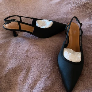 Svarta skor med taxklack - Aldrig använda svarta skor med taxklack och hälrem. Bred passform. Neopren på ovansidan och glansig klack. Klackhöjd: 5cm Storlek: 38 Köpta från Asos men aldrig använda!