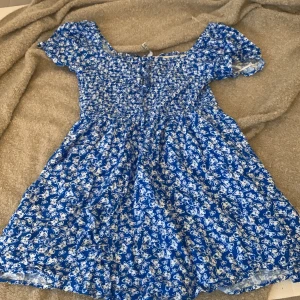Blå klänning med fjärilsmönster - En blå klänning med vita fjärilar. Klänningen har korta ärmar och en smockad överdel med knytband vid halsen. Den är kort och har en lös passform.