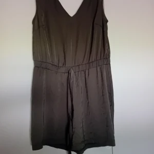 Brun klänning - En olivfärgad  shortsdress med V-ringning och ärmlös design. Klänningen har en enkel och elegant stil med en elastisk midja som kan knytas åt för en bekväm passform.