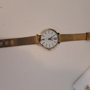 Guldfärgad klocka med vit urtavla - En elegant guldfärgad klocka med vit urtavla och svarta romerska siffror. Klockan har ett mesh-armband i guld och en klassisk spänne. Urtavlan är rund och har en minimalistisk design.
