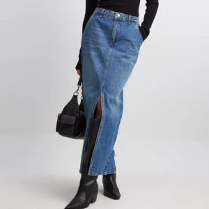 Blå jeanskjol - En lång blå jeanskjol med hög midja och slits framtill och baktill. Kjolen har en klassisk femficksdesign och stängs med dragkedja och knapp.