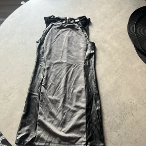 Svart och grå klänning från H&M - En elegant svart och grå klänning från H&M. Klänningen är ärmlös och har en dragkedja på baksidan. Den är tillverkad i ett glansigt material som ger en sofistikerad look.