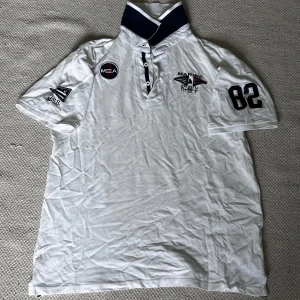 Vit pikétröja från Marine - En vit pikétröja från Marine Scandinavian Sports Wear med korta ärmar. Tröjan har flera tryck och broderier, inklusive siffran 82 på ärmen och olika logotyper på bröstet. Kragen är mörkblå med vita detaljer. Köpt secondhand och är i bra skick. Skriv för fler bilder eller frågor! 🤍💙❤️