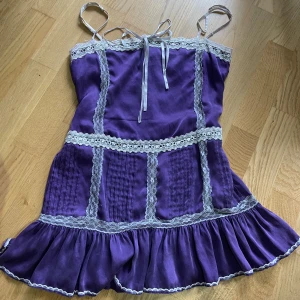 Lila klänning från Chick by Nicky Hilton - Linne från Chick by Nicky Hilton.Den har spetsdetaljer och smala axelband. Den har en volangkant längst ner och en knytning framtill.