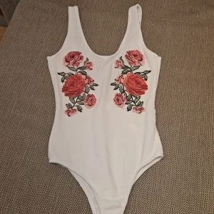 Vit baddräkt med blommigt mönster - En vit kroppsdrãkt med broderade röda och rosa blommor på framsidan. kroppsdräkten har breda axelband och en djup rygg. använd en gång bara. 100% ny. 