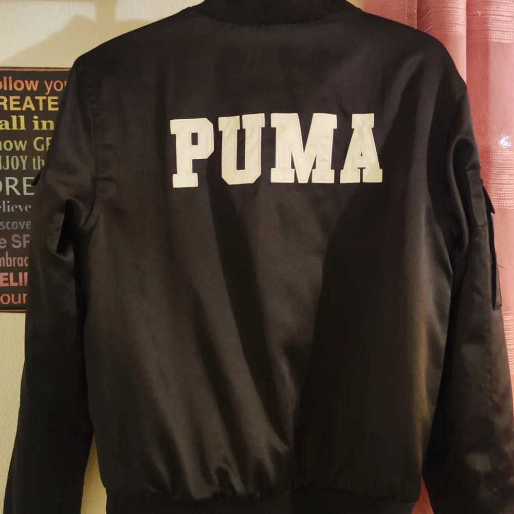 En svart bomberjacka från Puma med dragkedja framtill och vita Puma-logotyper på bröstet och ryggen. Jackan har långa ärmar och en ficka med knapp på ena ärmen.. Jackor.