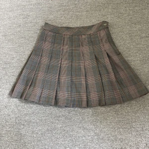 Rutig kjol från H&M - En rutig kjol från H&M tillverkad av 98% återvunnen polyester. Kjolen har ett rutigt mönster i grått, rött och beige med veckade detaljer och en knapp i midjan. Den är oanvänd och har lappen kvar