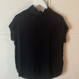 Svart topp från By Malene Birger - En elegant svart blus i silke från By Malene Birger med korta ärmar och en hög krage. Minimalt använd. Blusen har en dragkedja på baksidan.