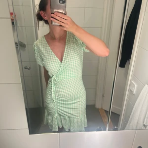 Grön rutig klänning från Gina Tricot - En grön rutig klänning från Gina Tricot med korta ärmar och omlott-design. Klänningen har en knytning i midjan och volangdetaljer vid fållen.