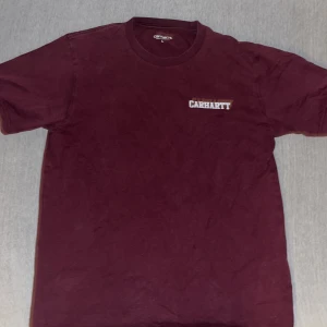 Vinröd t-shirt från Carhartt - En vinröd t-shirt från Carhartt med korta ärmar och rund halsringning. T-shirten har Carhartt-logotypen tryckt i vitt på bröstet.