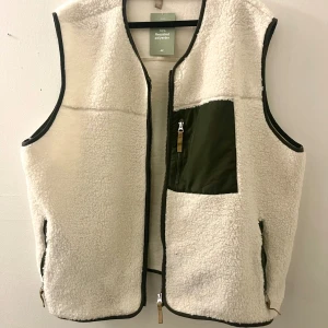 TEDDYVÄST  - Vit teddyväst med gröna detaljer, rundad hals med dragkedja. Grön ficka med dragkedja fram på bröstet. Strl. XXL normal i storleken, med utrymme för en hoodie under. Aldrig använd, prislappen är kvar, den är endast testad.