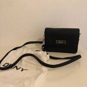 DKNY crossbody väska - En superfint, helt oanvänd väska från DKNY. Köpt i Köpenhamn för några år sedan för runt 2000 kr! Axelbandet är justerbart, men även avtagbart. Den kan skickas på posten, men då får köparen stå för frakten! Kan alltid annars mötas upp i Malmö :)