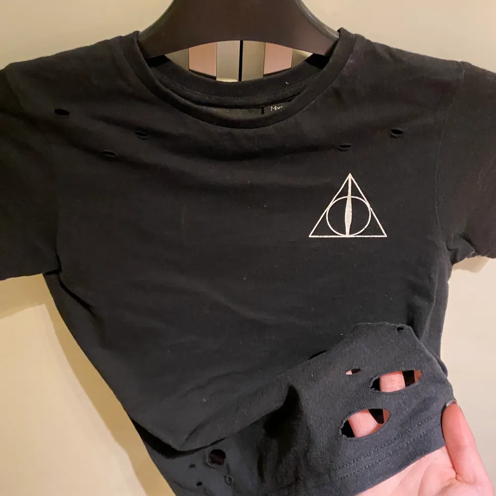 Svart t-shirt med dödsrelikerna från Harry Potter tryck på framsida och baksida. Även tillgorda hål i kanten på tröjan samt lite vid nyckelbenen. (Hålen är en del av designen) Köpt i london, fin tröja i bra skick säljer pga att den var för liten🥰 . T-shirts.