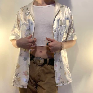 Vintage vit glansig pyjamaströja blommor - Vitt vintage oversized glansig kortärmad pyjamaströja i siden imitation med en söt krage, ficka på vänster bröst, knappar av samma tyg som resten av tröjan och det coolaste blommiga vattenfärg mönstret 🌸 Denna tröjan är så versatile då man kan använda den som en faktisk pyjamaströja, som en skön oversized skjorta som man slänger över en baby tee och ett par ljusa byxor eller som en mer fancy topp med ett par kostymbyxor och gogo boots! Kvalitén är otrolig och tröjan är så så skön!
