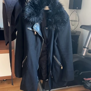 Svart Nelly kappa med fake pälskrage  - Svart halv lång kappa från Nelly, väl använd men fortfarande i gott skick!!! Skinn detaljer, dragkedjor och pälskrage av falsk päls. Storlek 38